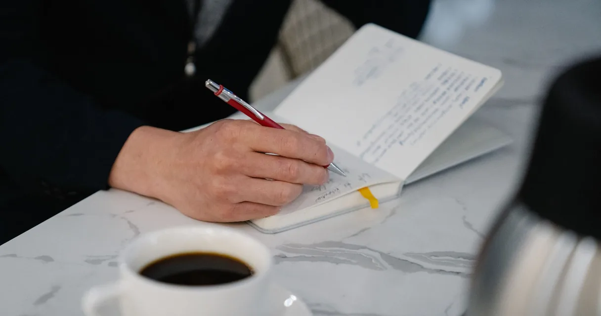 Mão escrevendo anotações em um caderno ao lado de uma xícara de café, rotina de hábitos para parar de beber