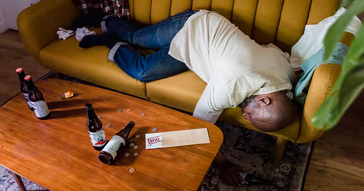 Homem desmaiado no sofá com garrafas de cerveja caídas na mesa, cena de quem bebe muito